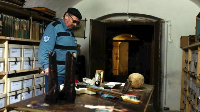 Ein Mann mit Beret steht im Keller und blickt auf einen Schreibtisch auf dem viele Erinnerungsstücke und Bilder stehen. Ein Mann mit Beret steht im Keller und blickt auf einen Schreibtisch auf dem viele Erinnerungsstücke und Bilder stehen.