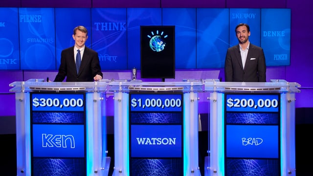 Zwei Männer und ein Computer stehen hinter den Kandidatenpulten der US-Quizshow Jeopardy! Zwei Männer und ein Computer stehen hinter den Kandidatenpulten der US-Quizshow Jeopardy!