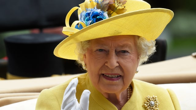 Queen Nahe mit gelbem Hut und gelber Kleidung, winkend und leicht lächelnd