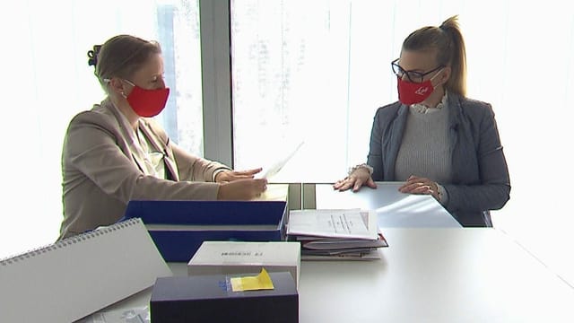 Zwei Frauen in Anzügen und mit roten Masken sitzen an einem Tisch. Auf dem Tisch liegen Papier und Ordner. Zwei Frauen in Anzügen und mit roten Masken sitzen an einem Tisch. Auf dem Tisch liegen Papier und Ordner.