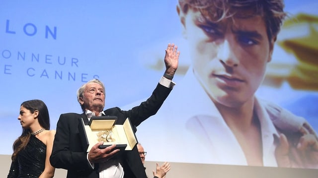 Alain Delon steht auf einer Bühne und hält die Goldene Palme in der Hand. Hinter ihm ist ein grosses Porträt von ihm in jungen Jahren. Alain Delon steht auf einer Bühne und hält die Goldene Palme in der Hand. Hinter ihm ist ein grosses Porträt von ihm in jungen Jahren.