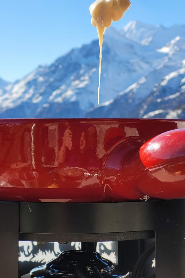 Ein Fonduecaquelon und ein Brocken Fondue mit Käse vor der Berglandschaft.