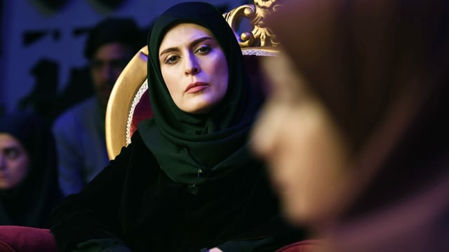 Filmszene: Eine Frau mit Kopftuch sitzt in einem pompösen Sessel und schaut ernst auf eine jüngere Frau im Vordergrund, die man nicht richtig erkennt. Filmszene: Eine Frau mit Kopftuch sitzt in einem pompösen Sessel und schaut ernst auf eine jüngere Frau im Vordergrund, die man nicht richtig erkennt.