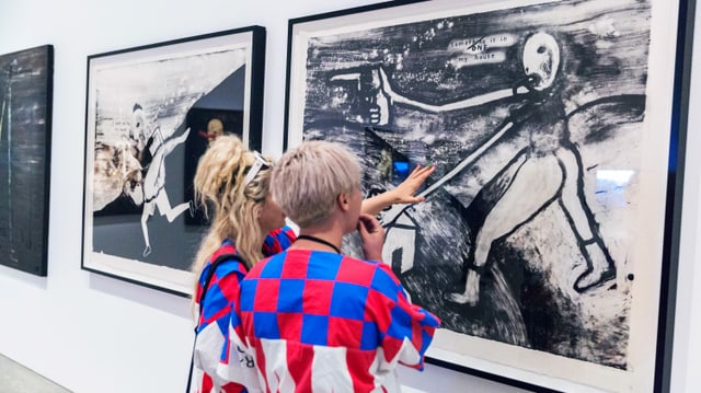 Zwei junge Frauen schauen sich in einem Museum ein Bild an. Es ist mit schwarzer Farbe auf einen weissen Hintergrund gemalt und zeigt eine abstrakte Figur mit einer Pistole in der Hand, die sie nach vorne richtet. Darunter ist ein simpel gezeichnetes Haus zu sehen. Über der Figur steht: "Someone is in my house." Zwei junge Frauen schauen sich in einem Museum ein Bild an. Es ist mit schwarzer Farbe auf einen weissen Hintergrund gemalt und zeigt eine abstrakte Figur mit einer Pistole in der Hand, die sie nach vorne richtet. Darunter ist ein simpel gezeichnetes Haus zu sehen. Über der Figur steht: "Someone is in my house."