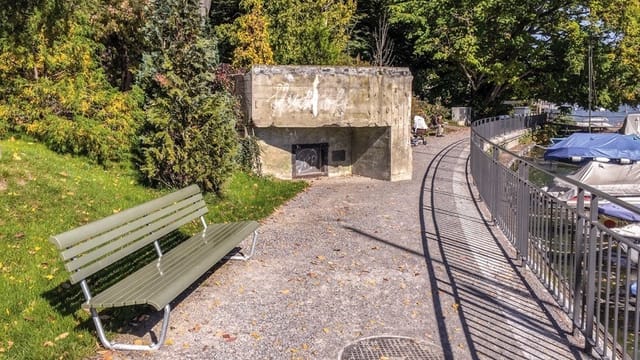 Ein Bunker an der Seepromenade, im Vordergrund ein Kiesweg, links eine SItzbank, dahinter der Bunker und Bäume. Ein Bunker an der Seepromenade, im Vordergrund ein Kiesweg, links eine SItzbank, dahinter der Bunker und Bäume.