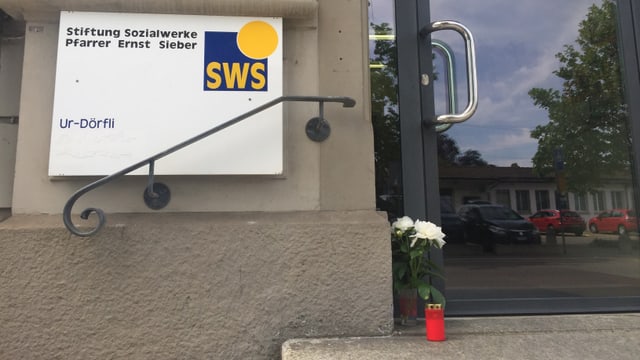 Eingangstüre zum Ur-Dörfli, am Boden stehen eine rote Kerze und zwei weisse Rosen. Eingangstüre zum Ur-Dörfli, am Boden stehen eine rote Kerze und zwei weisse Rosen.