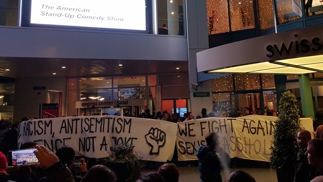 Menschenmenge vor einem Gebäude, einige halten Plakate. Aufschrift: "Racism, Antisemitism... not a joke." Menschenmenge vor einem Gebäude, einige halten Plakate. Aufschrift: "Racism, Antisemitism... not a joke."