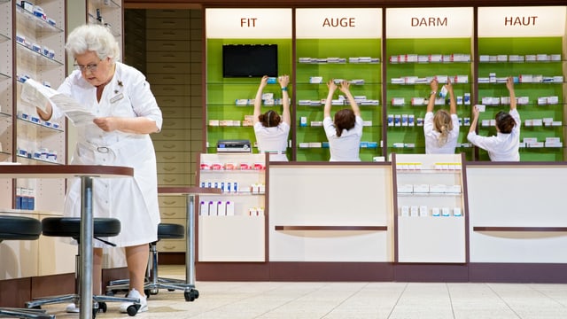 5 Frauen in Kitteln in einer Apotheke, eine liest eine Liste, die anderen füllen Regale auf. 5 Frauen in Kitteln in einer Apotheke, eine liest eine Liste, die anderen füllen Regale auf.