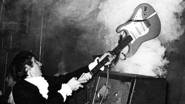 Ein junger Mann im schwarzen Anzug schlägt seine Gitarre auf einen rauchenden Verstärker. Ein junger Mann im schwarzen Anzug schlägt seine Gitarre auf einen rauchenden Verstärker.