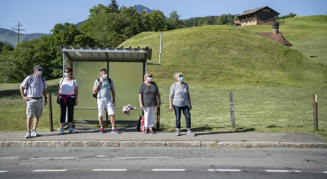 Personen warten in ländlicher Umgebung auf einen Bus – alle tragen Hygienemasken. Personen warten in ländlicher Umgebung auf einen Bus – alle tragen Hygienemasken.