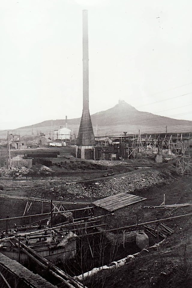 Schwarzweiss-Bild einer verlassenen Grabungsstätte mit Hütten und einem grossen Kamin. 