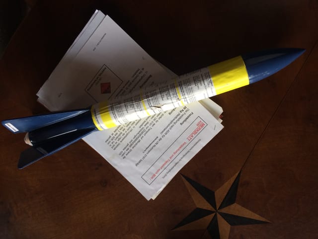 Blau-Gelbe Rakette liegt auf Blättern mit Aufschrift "Merkblatt", darunter Holztisch Blau-Gelbe Rakette liegt auf Blättern mit Aufschrift "Merkblatt", darunter Holztisch