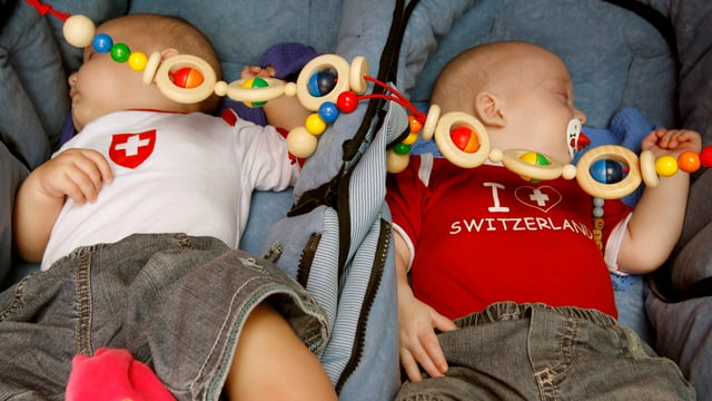 Zwei schlafende Babys in einem Doppelkinderwagen, beide tragen T-Shirts mit Schweizerkreuz.