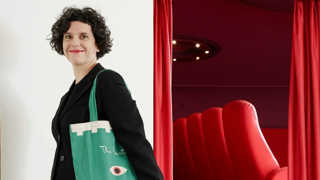 Eine jüngere Frau steht vor dem Eingang zu einem Kinosaal, der mit roten Sesseln bestückt ist. Eine jüngere Frau steht vor dem Eingang zu einem Kinosaal, der mit roten Sesseln bestückt ist.