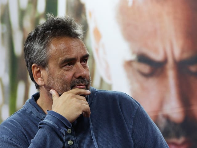 Luc Besson hält seine Hand an sein Kinn. Im Hintergrund ist ein Plakat von ihm. Luc Besson hält seine Hand an sein Kinn. Im Hintergrund ist ein Plakat von ihm.