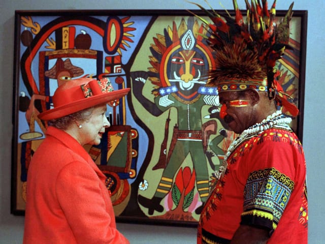 In der linken Bildhälfte sieht man die Queen mit einem roten Mantel und einem roten Hut. Recht davon steht ein Künstler aus Papua-Neuguinea mit einem traditionellen Hemd und einem Federhut – ebenfalls in rot. Im Hintergrund sieht man ein Bild des Künstlers.