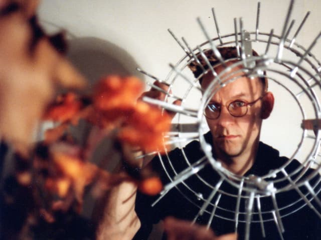 Ein Amnn mit Brille schaut durch ein rundes Objekt aus Metall in die Kamera. Ein Amnn mit Brille schaut durch ein rundes Objekt aus Metall in die Kamera.