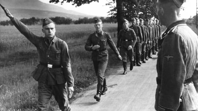 Junge Männer marschieren in Uniform auf einer Landstrasse, einer zeigt den Hitlergruss. Junge Männer marschieren in Uniform auf einer Landstrasse, einer zeigt den Hitlergruss.