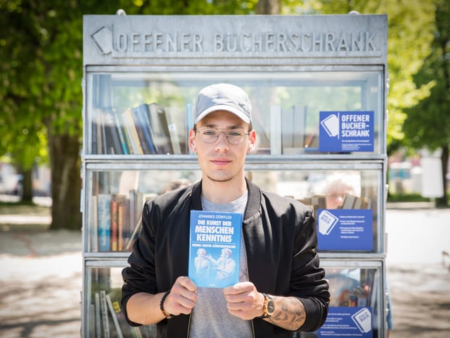 Junger Mann mit Baseballkape steht vor einem Bücherregal, das draussen an einem Wegrand steht.