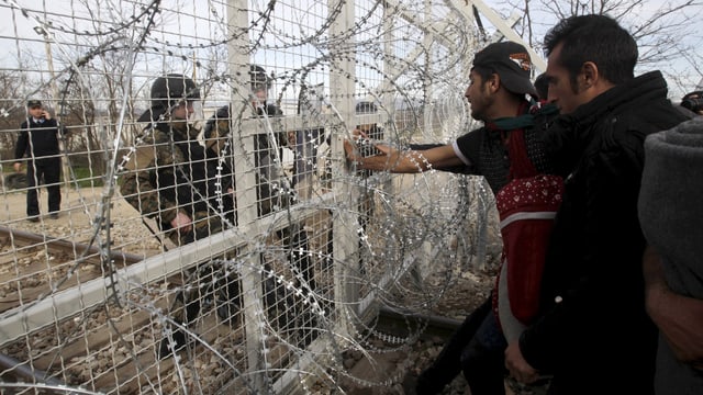 Flüchtlinge blicken durch einen Zaun hindurch zwei Polizisten an. Flüchtlinge blicken durch einen Zaun hindurch zwei Polizisten an.