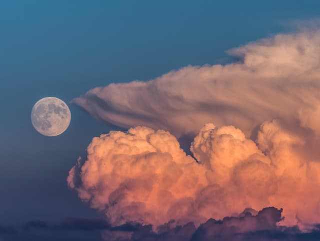 In der rechten Bildhälfte steht ein Gewitterturm im Sonnenuntergang. Links liegt der Mond.