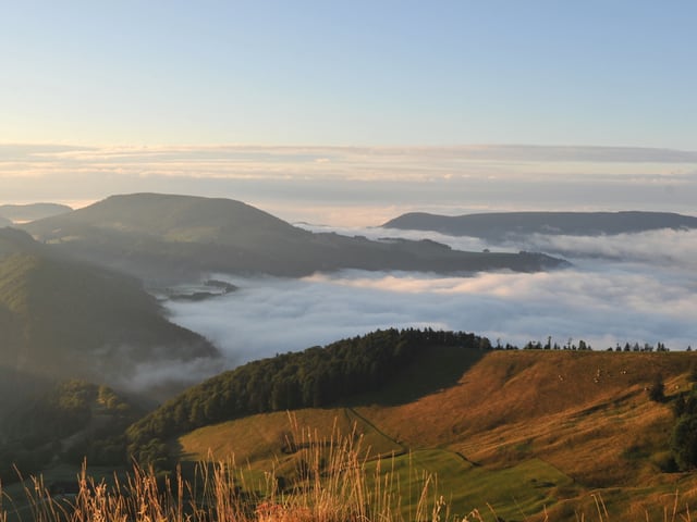 Die Hügel mit Wälder und Wiese werden von der Morgensonne bereits beschienen. Zwischen den Hügel hat es Nebel. Der Himmel ist abgesehen von wenigen Wolkenfeldern blau.