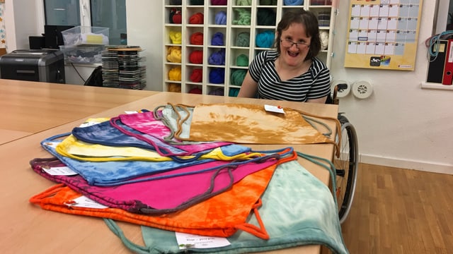 Eine Frau im Rollstuhl zeigt batik-gefärbte Baumwolltaschen in der Werkstatt des Wagerenhofs. Eine Frau im Rollstuhl zeigt batik-gefärbte Baumwolltaschen in der Werkstatt des Wagerenhofs.