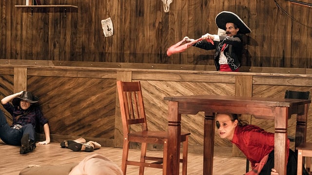 Drei Schauspieler in einem Westernsaloon. Einer sitzt am Boden, eine Frau unter einem Tisch und ein Mann hinter der Bar. Drei Schauspieler in einem Westernsaloon. Einer sitzt am Boden, eine Frau unter einem Tisch und ein Mann hinter der Bar.