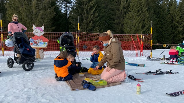 Frau mit Kindern auf Decke. Sie sitzen im Schnee und essen Picknick.