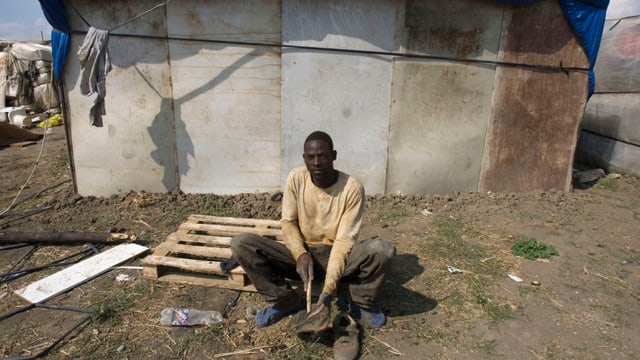 Ein afrikanischer Mann sitzt vor einer baufälligen Hütte auf einem Feld. Ein afrikanischer Mann sitzt vor einer baufälligen Hütte auf einem Feld.