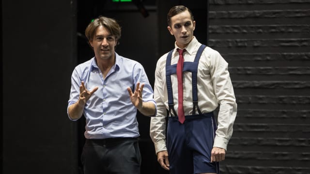 Zwei Männer stehen auf der Bühne. Der Mann rechts in kurzen Hosen, Hosenträgern und einer roten Krawatte. Zwei Männer stehen auf der Bühne. Der Mann rechts in kurzen Hosen, Hosenträgern und einer roten Krawatte.
