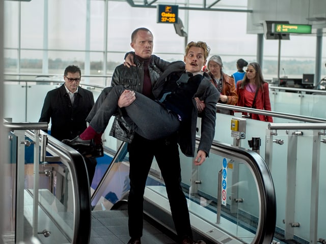 Jock trägt Mortdecai über die Rolltreppe im Flughafen. Jock trägt Mortdecai über die Rolltreppe im Flughafen.