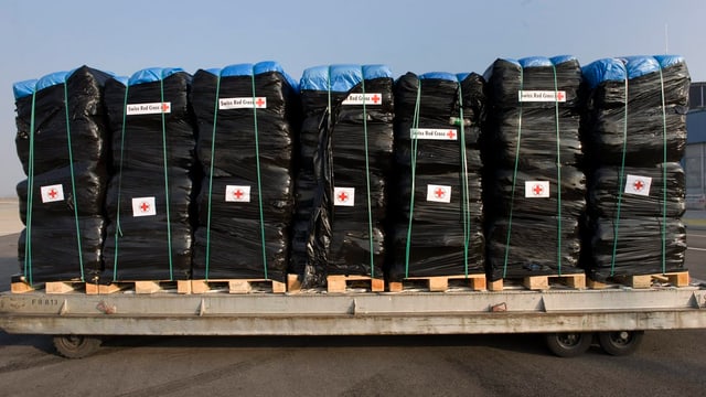 Eine Palette voller in blauen und schwarzen Plastik gehüllte Hilfsgüter mit Swiss-Red-Cross-Klebern.