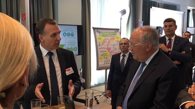 Bundesrat Schneider-Ammann steht im Anzug in einem Raum und horcht einer Person, die ihm etwas erklärt. Bundesrat Schneider-Ammann steht im Anzug in einem Raum und horcht einer Person, die ihm etwas erklärt.