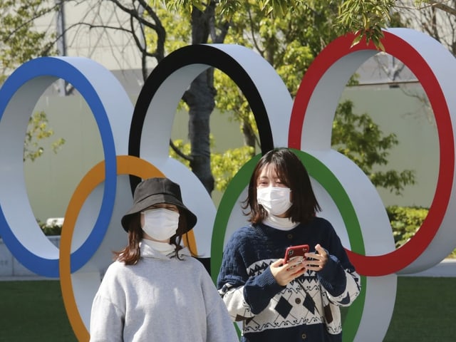 Frauen mit Masken vor den olympischen Ringen.