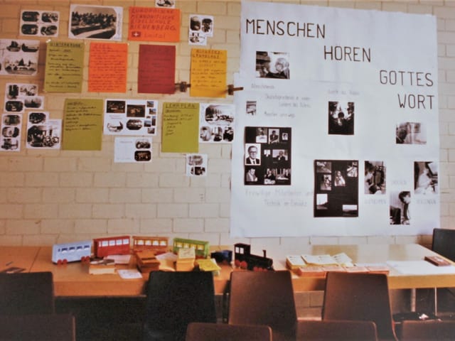 Man sieht eine Backsteinwand, vor welcher zwei Tische angeordnet sind. An der Backsteinwand hängen viele farbige Plakate und Bilder. Neben den farbigen Plakaten hängt ein grosses Bild mit der Aufschrift "Menschen hören Gottes Wort". Man sieht eine Backsteinwand, vor welcher zwei Tische angeordnet sind. An der Backsteinwand hängen viele farbige Plakate und Bilder. Neben den farbigen Plakaten hängt ein grosses Bild mit der Aufschrift "Menschen hören Gottes Wort".