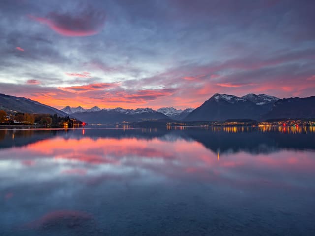 See in lila bis rosa Morgenstimmung mit Bergen an den Ufern und leicht bewölktem Himmel