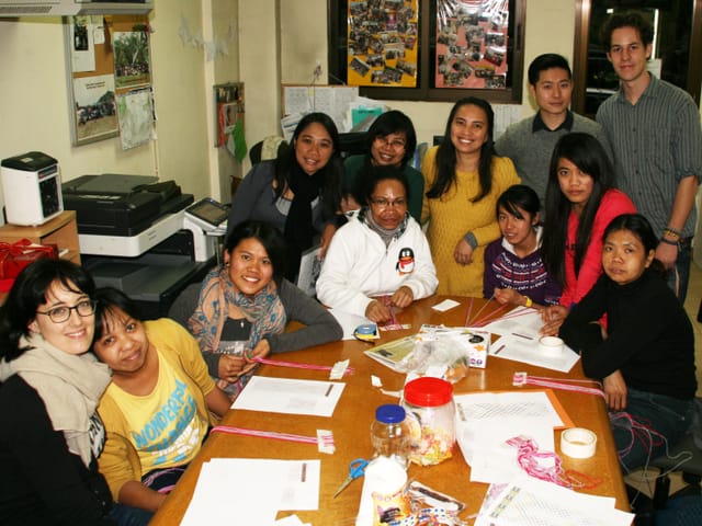 Gruppenbild verschiedener Indonesierinnen und zwei Helfern, um einen Tisch herum gruppiert. Gruppenbild verschiedener Indonesierinnen und zwei Helfern, um einen Tisch herum gruppiert.