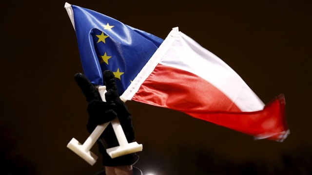 Eoine Hand in einem schwarzen Handschuh streckt vor dunklem Himmel eine EU- und Polenflagge in die Höhe. Eoine Hand in einem schwarzen Handschuh streckt vor dunklem Himmel eine EU- und Polenflagge in die Höhe.