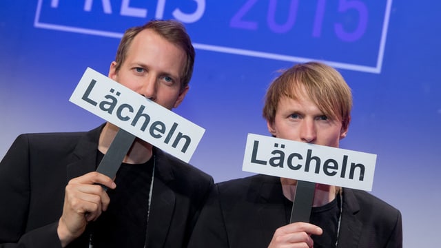 Zwei Männer halten Schilder hoch, auf denen «Lächeln» steht. Zwei Männer halten Schilder hoch, auf denen «Lächeln» steht.