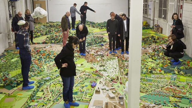 buntes Stadtmodell in einem grossen Raum, darauf stehen Leute buntes Stadtmodell in einem grossen Raum, darauf stehen Leute