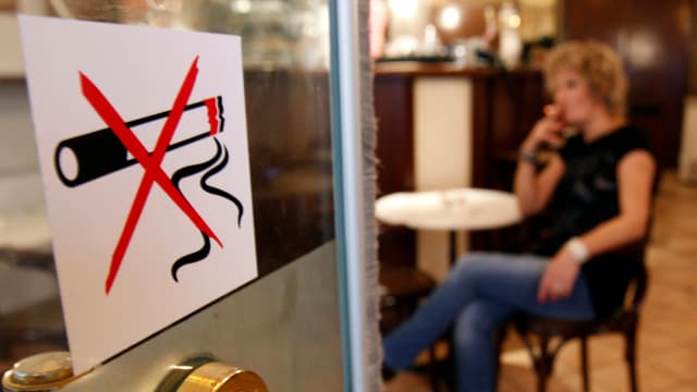 An der EingangstÃ¼r eines Lokals klebt ein Rauchverbotszeichen. Im Innern des CafÃ©s ist eine Frau zu sehen, die eine Zigarette raucht.