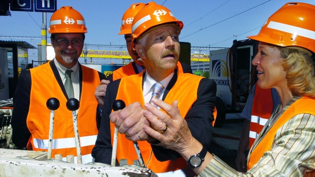 Drei Männer und eine Frau mit orangefarbenen Helmen stehen an Schalthebeln. Drei Männer und eine Frau mit orangefarbenen Helmen stehen an Schalthebeln.