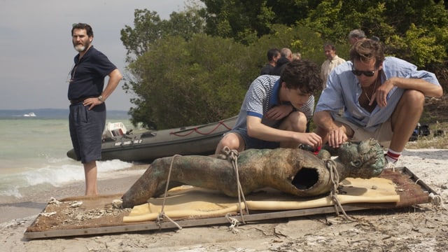 Am Strand liegt eine Metallfigur, zwei Männer beugen sich darüber und einer steht daneben. Am Strand liegt eine Metallfigur, zwei Männer beugen sich darüber und einer steht daneben.