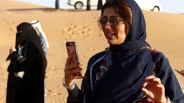 Symbolbild: Frau ohne Schleier mit Handy in der Wüste, dahinter Frau mit Schleier. Symbolbild: Frau ohne Schleier mit Handy in der Wüste, dahinter Frau mit Schleier.