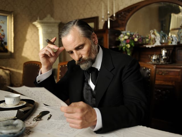 Filmszene: Freud im Anzug mit Zigarre am Tisch, einen Brief lesend. Filmszene: Freud im Anzug mit Zigarre am Tisch, einen Brief lesend.