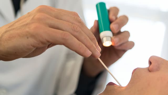 Hand mit Holzstäbchen und Hand mit Taschenlampe vor einem geöffneten Mund eines liegenden Patienten Hand mit Holzstäbchen und Hand mit Taschenlampe vor einem geöffneten Mund eines liegenden Patienten