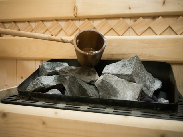 In einer Sauna steht ein kleines Metallbecken mit Steinen. Über diese Steine wird mit einer Holzkelle Wasser geleert. In einer Sauna steht ein kleines Metallbecken mit Steinen. Über diese Steine wird mit einer Holzkelle Wasser geleert.