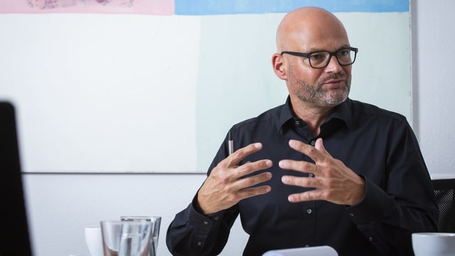 Ein Mann mit Brille spricht und gestikuliert vor einem pastellfarbenen Gemälde. Ein Mann mit Brille spricht und gestikuliert vor einem pastellfarbenen Gemälde.