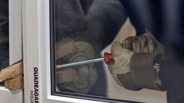 Zwei Hände mit Handschuhen versuchen mit Holzstücken und einem Schraubenzieher ein Fenster aufzubrechen. Zwei Hände mit Handschuhen versuchen mit Holzstücken und einem Schraubenzieher ein Fenster aufzubrechen.